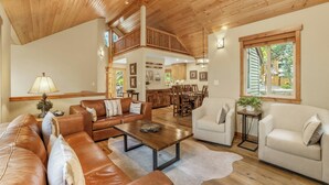 A spacious living area with a high ceiling, wooden flooring, and a cozy seating arrangement including a brown leather sofa, a white armchair, and a wooden coffee table.