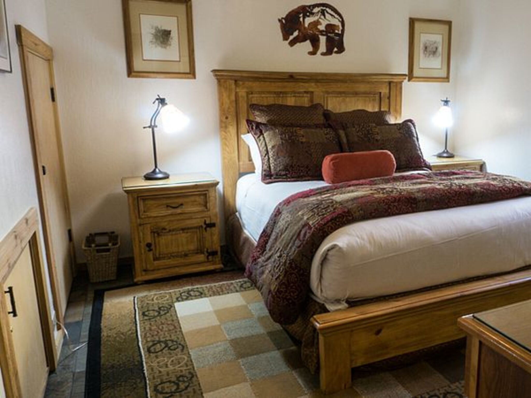 A bedroom with a wooden bed frame, two bedside tables with lamps, a patterned rug, and a wall-mounted bear decoration.