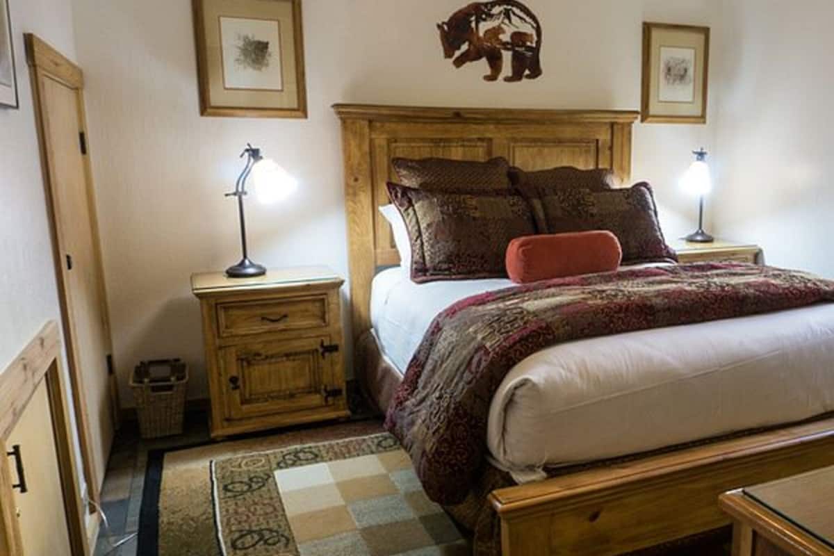 Un dormitorio con estructura de cama de madera, dos mesitas de noche con lámparas, un tapete estampado y un adorno de oso colgado en la pared.