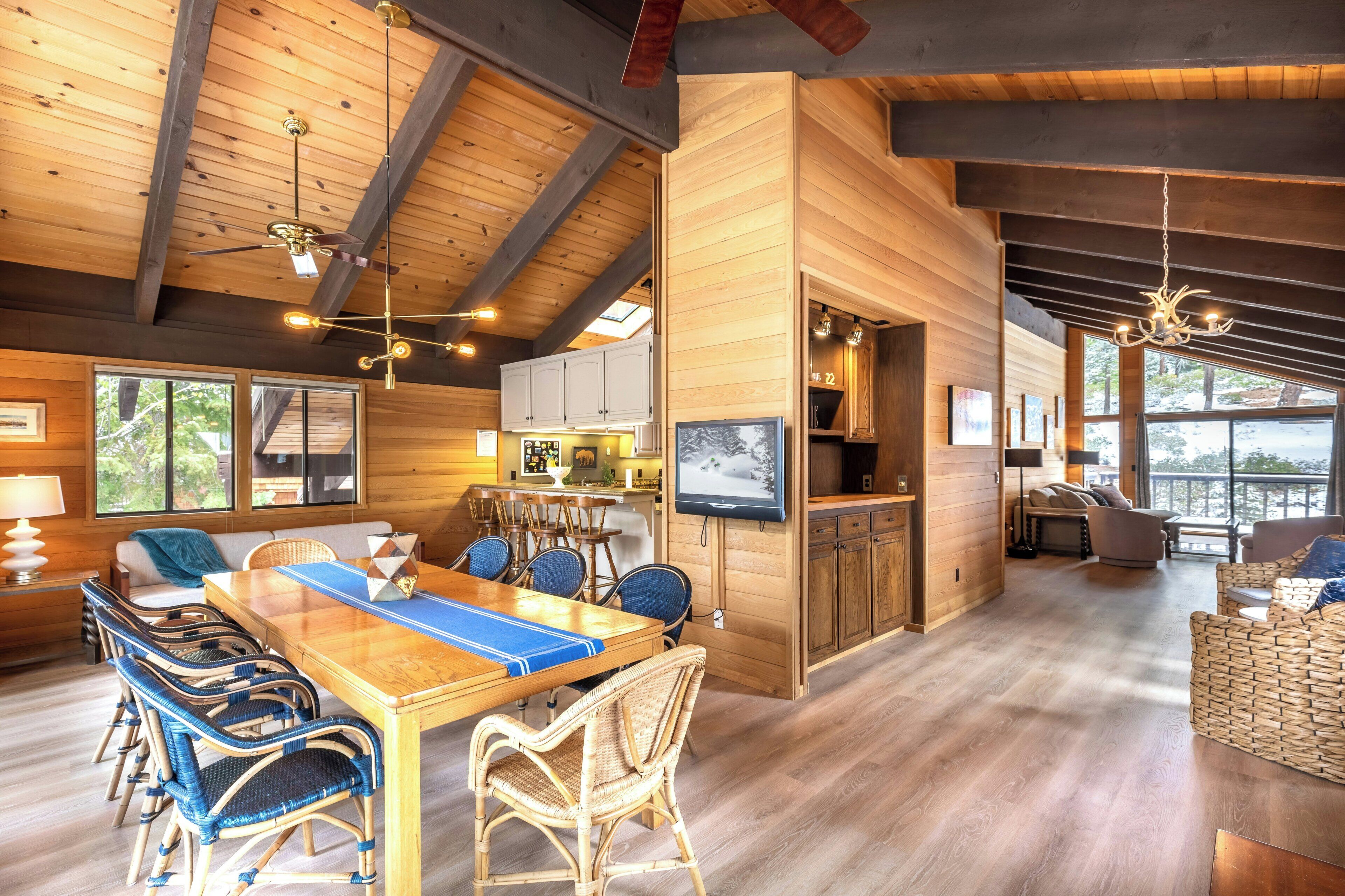 Ein geräumiges Zimmer mit Esstisch aus Holz, Korbstühlen, einem Fernseher und einem großen Fenster mit Blick ins Freie.
