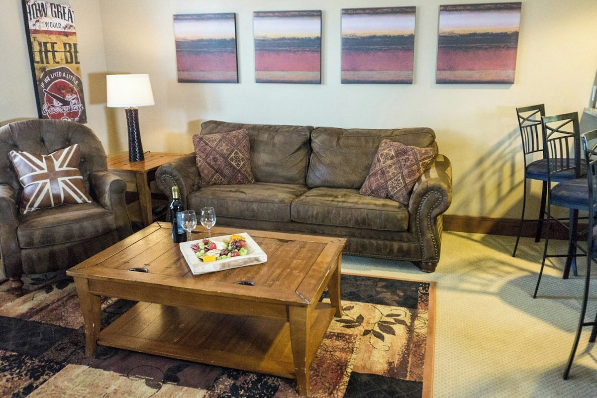 Una sala de estar con un sofá de cuero marrón, una mesa de centro de madera con una bandeja de frutas, un tapete estampado y una pared con tres cuadros de atardecer.