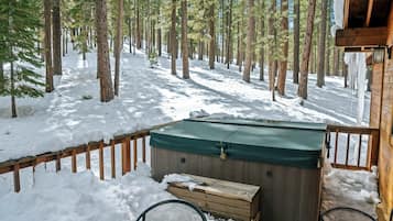 A wooden deck with a hot tub, surrounded by snow-covered ground and tall pine trees.
