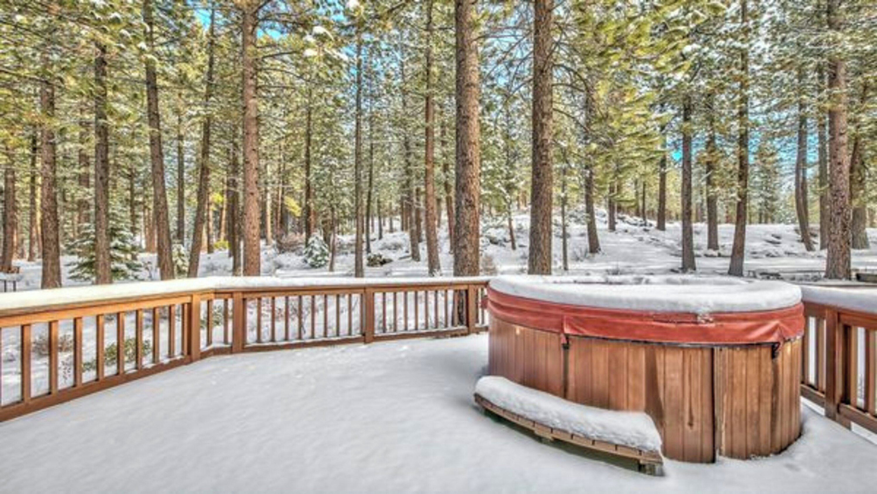 Una terraza cubierta de nieve con un jacuzzi y barandillas de madera, rodeada de altos árboles.