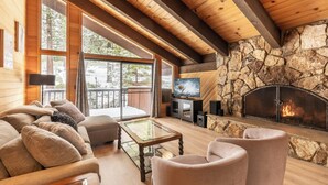 A cozy living room with a stone fireplace, wooden beams, and large windows overlooking a snowy deck.