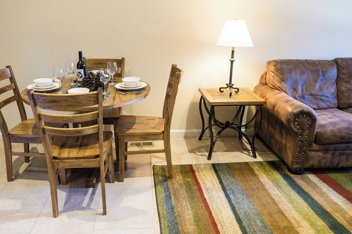 Un comedor con una mesa de madera, sillas y una lámpara. Hay un sofá y una alfombra a rayas en la sala.