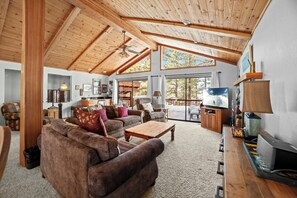 A spacious living room with a high ceiling, wooden beams, large windows, a fireplace, and a television. - Northstar Resort by Vail Resorts (Truckee)
