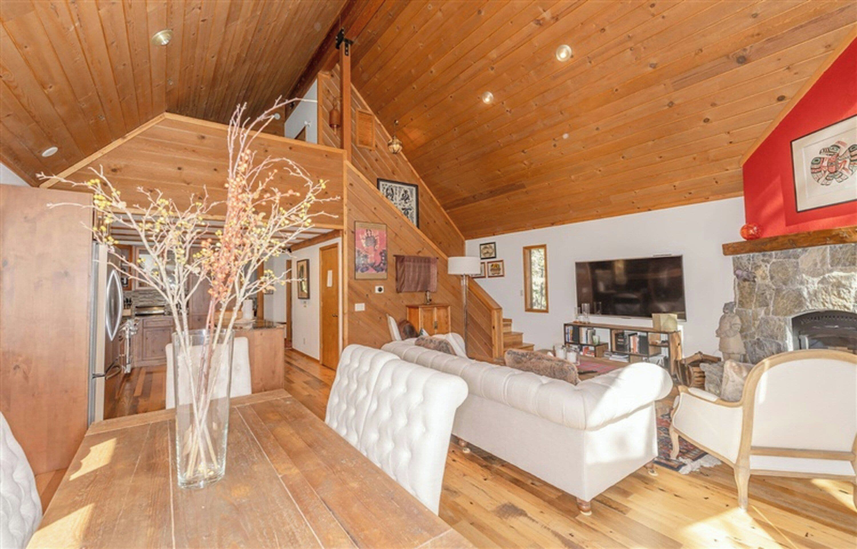 A cozy living room with a wooden ceiling, a stone fireplace, a white sofa, and a dining table with a vase of branches.