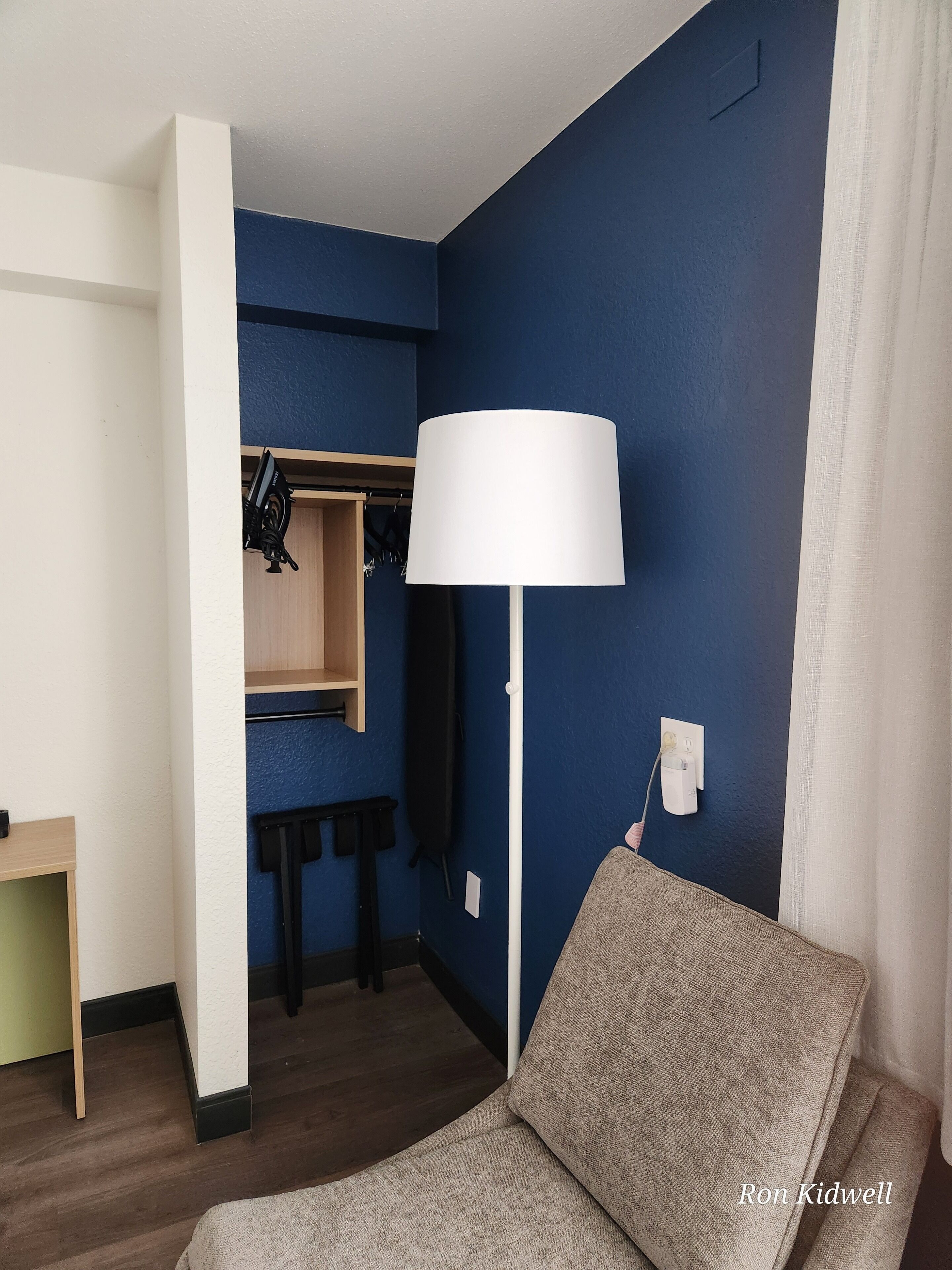 A hotel room with a blue accent wall, a white lamp, a grey sofa, and a wooden side table.