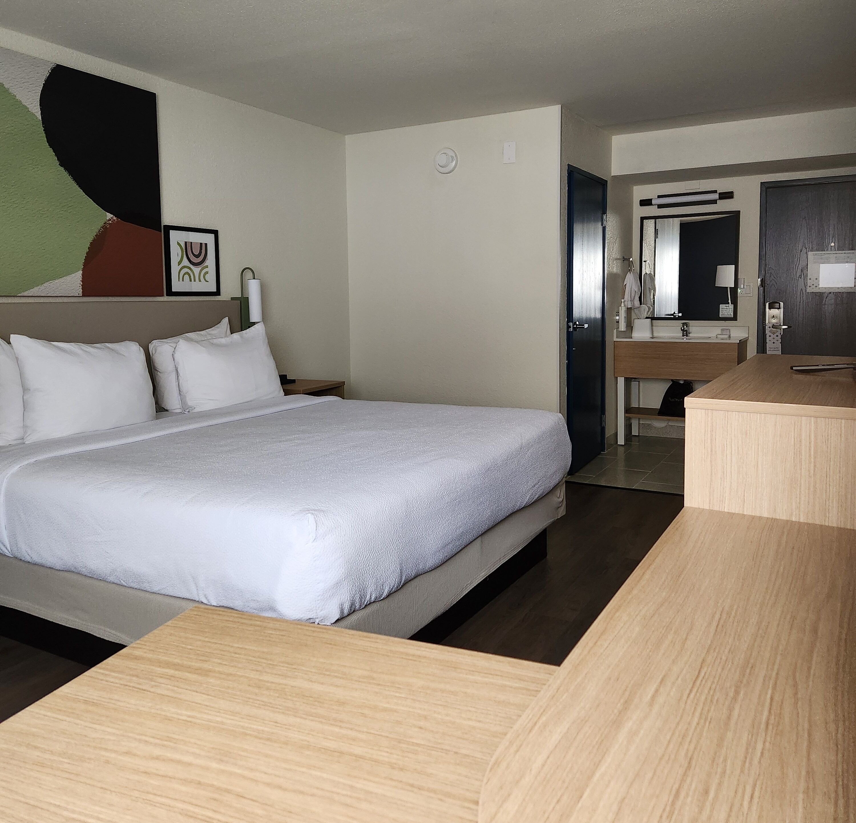 A hotel room with a large bed, a bedside table, a bathroom with a sink and mirror, and a wall-mounted air conditioning unit.