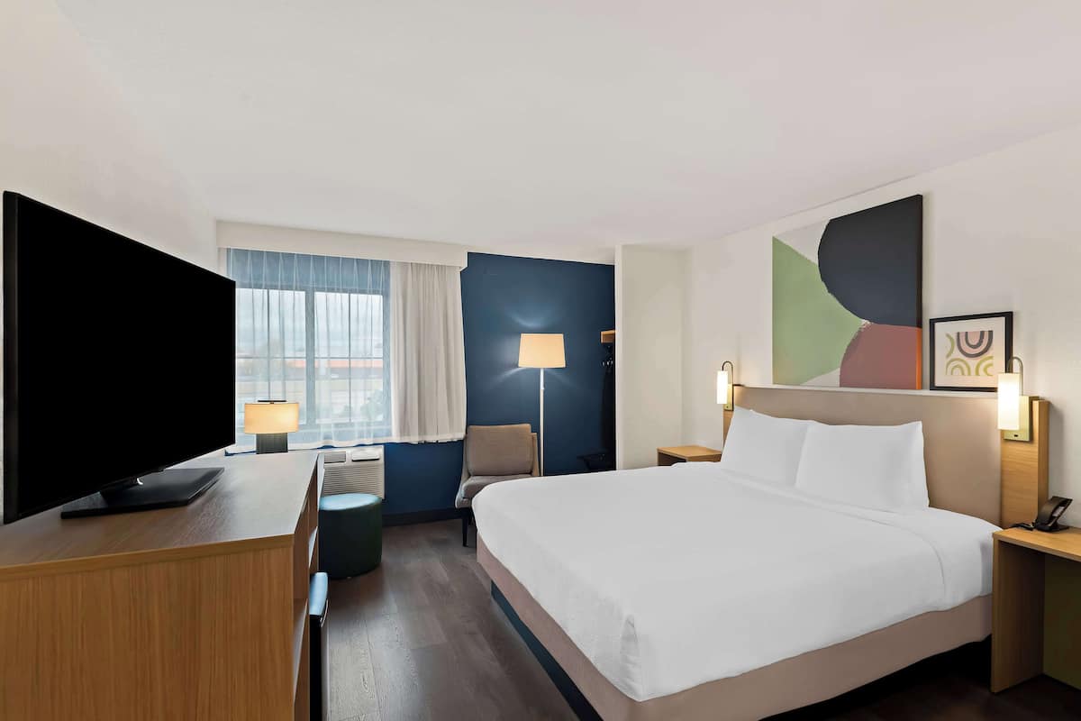 A hotel room with a large bed, a desk with a flat-screen TV, a chair, a lamp, and a window with curtains.