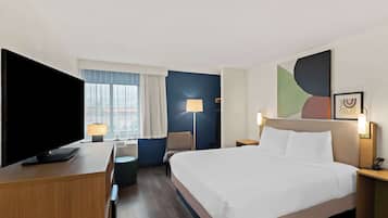 Habitación de hotel con una cama grande, un escritorio con televisor de pantalla plana, una silla, una lámpara y una ventana con cortinas.