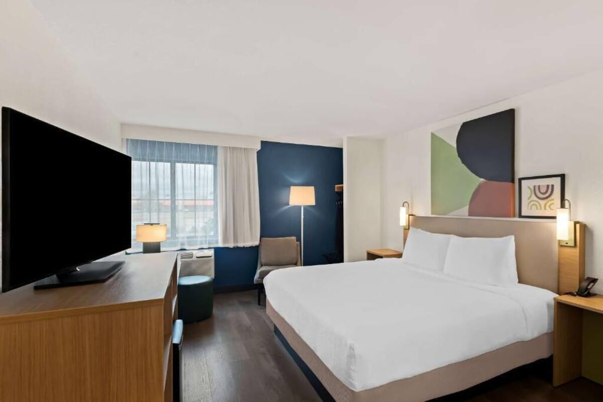 A hotel room with a large bed, a desk with a flat-screen TV, a chair, a lamp, and a window with curtains.