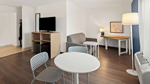 Una habitación de hotel moderna con televisión de pantalla plana, un sofá, una mesita con lámpara, una mesa redonda con sillas y un radiador.