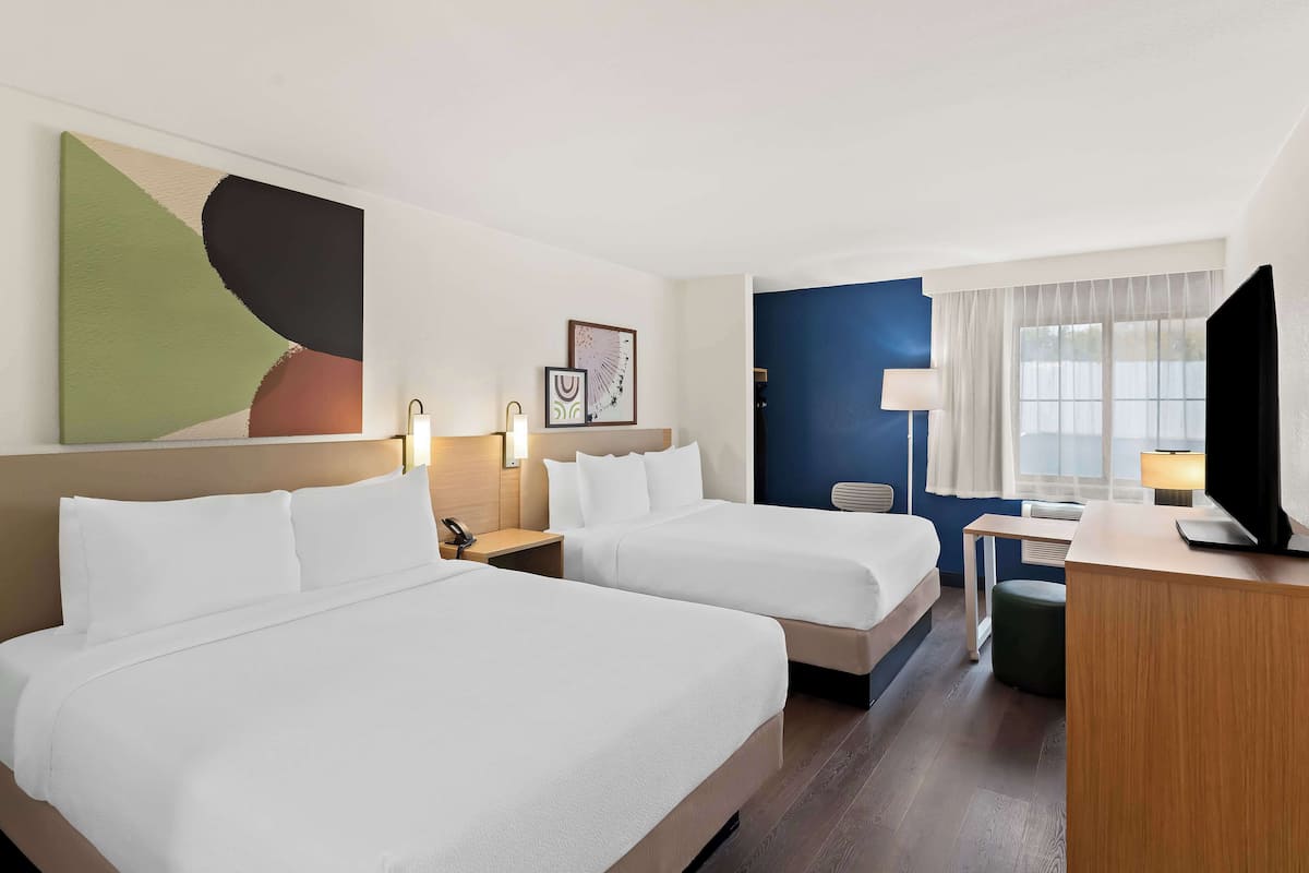 A hotel room with two beds, a desk, a chair, a TV, and a large abstract painting on the wall.