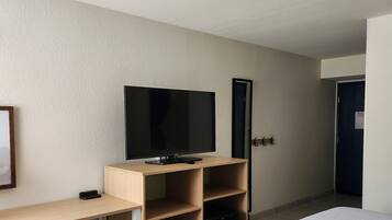 A hotel room with a bed, a desk with a television, a trash bin, and a door.