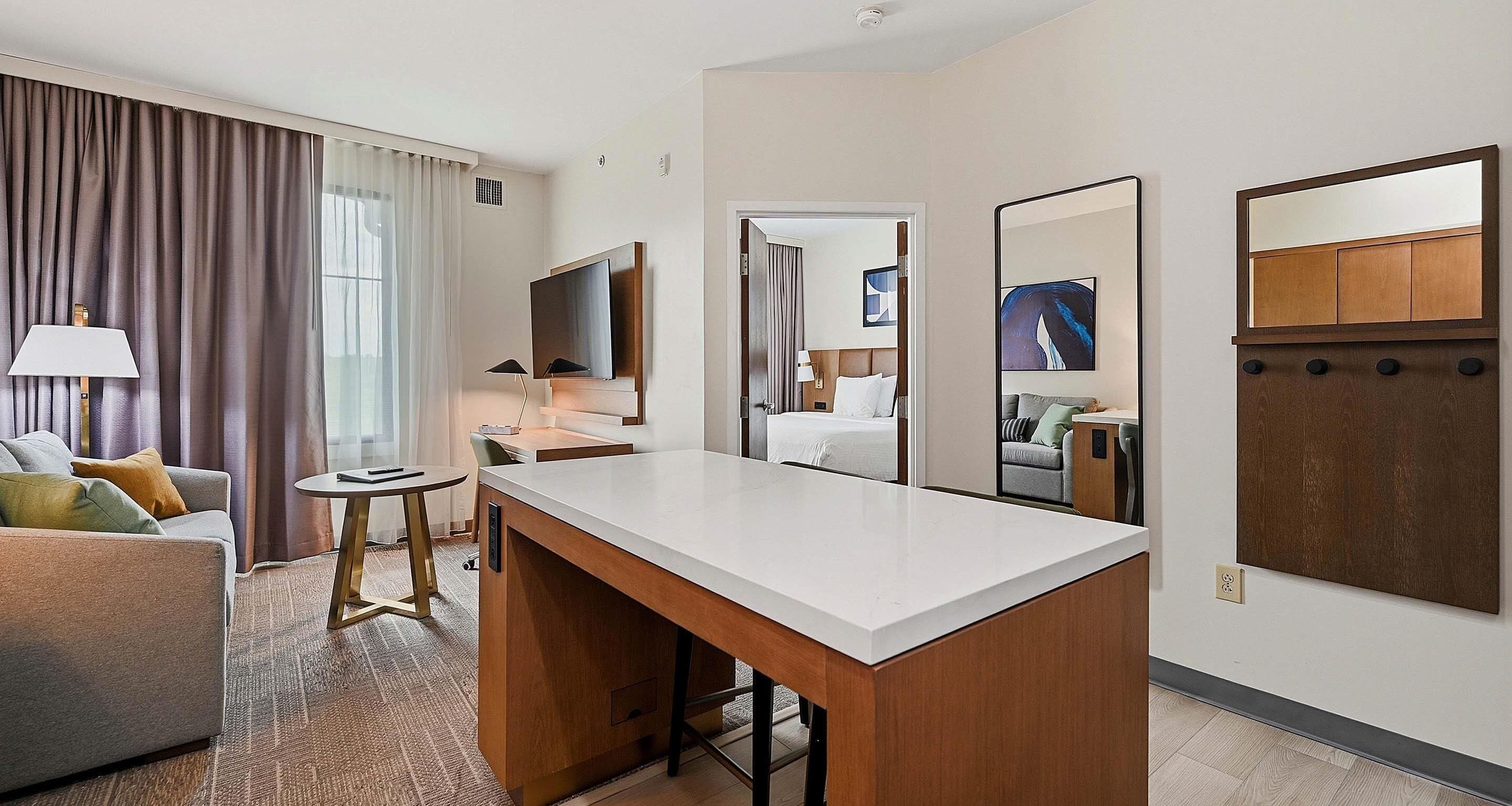 Habitación de hotel con escritorio, sofá, mesita, lámpara, televisor, espejo y obras de arte en la pared.