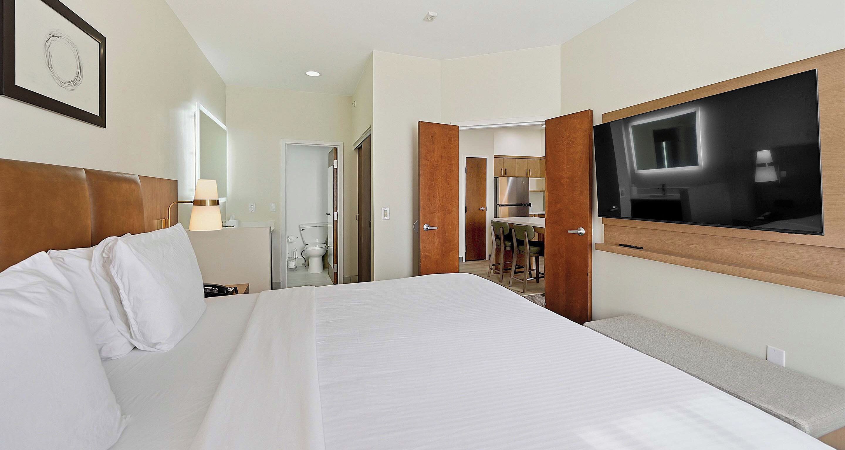 A hotel room with a large bed, a TV mounted on the wall, a bathroom with a toilet and sink, and a wooden headboard.