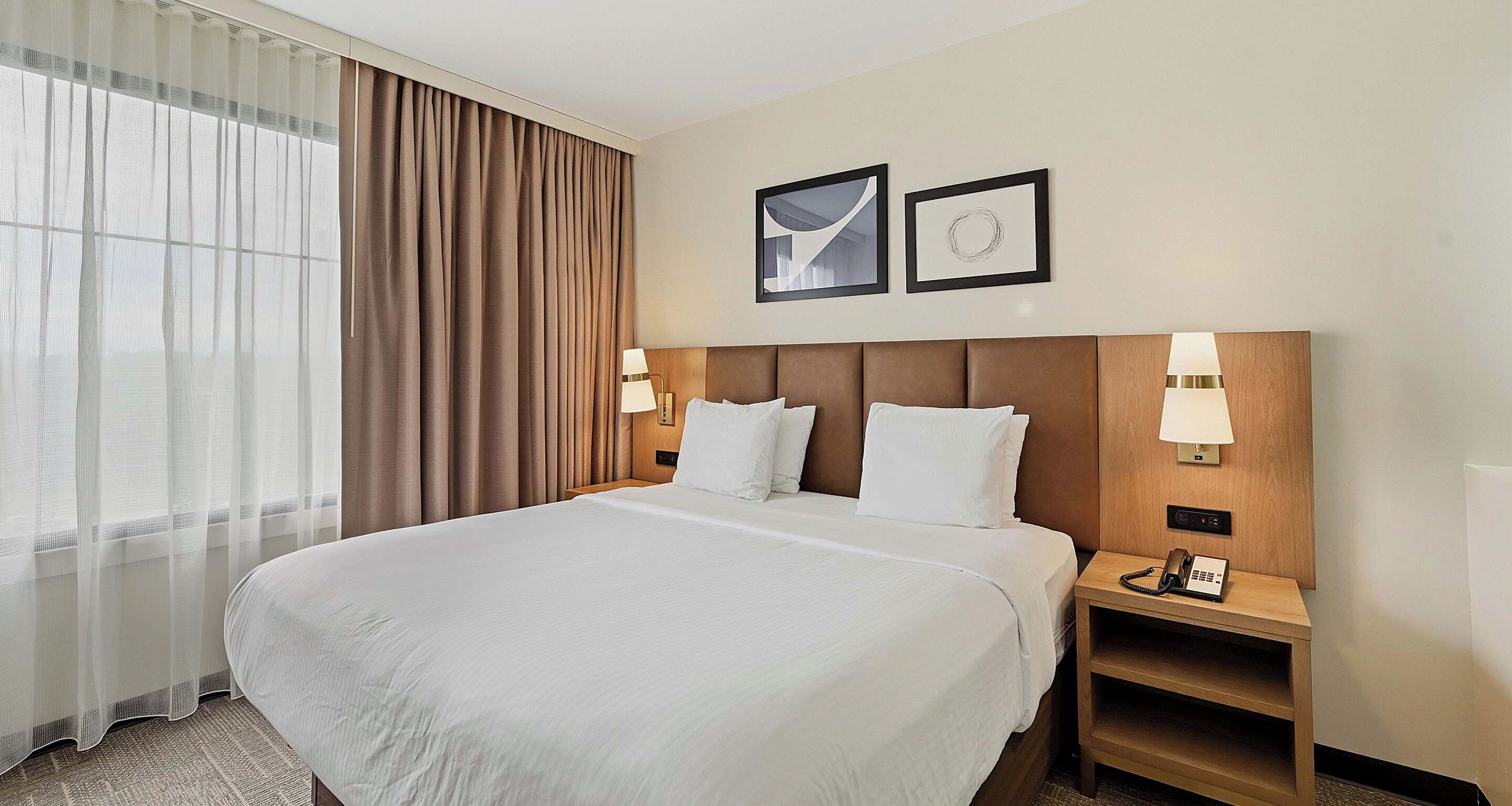 Een hotelkamer met een groot bed, twee bedlampjes, een nachtkastje met een telefoon en een raam met dunne gordijnen.