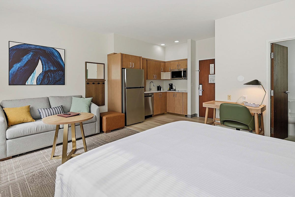 A hotel room with a bed, a sofa, a small table, a desk, a kitchen area, and a painting on the wall.