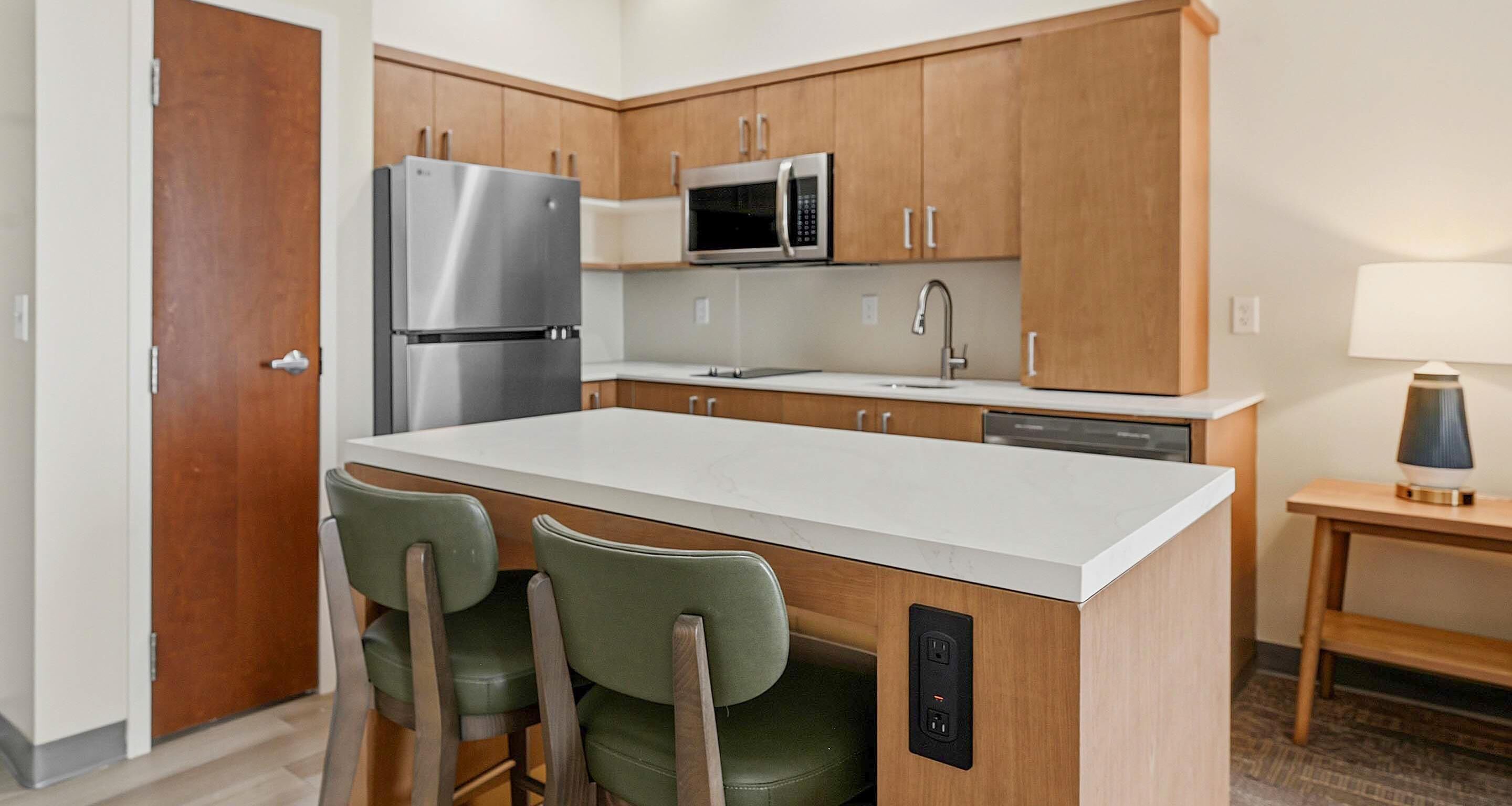 A modern kitchen with wooden cabinets, a stainless steel refrigerator, a microwave, a sink, and a bar stool.