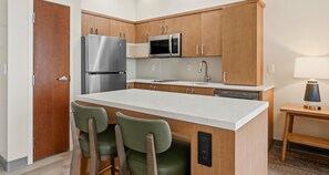 A modern kitchen with wooden cabinets, a stainless steel refrigerator, a microwave, a sink, and a bar stool. - Best Western Plus Hannaford Inn & Suites (Cincinnati)