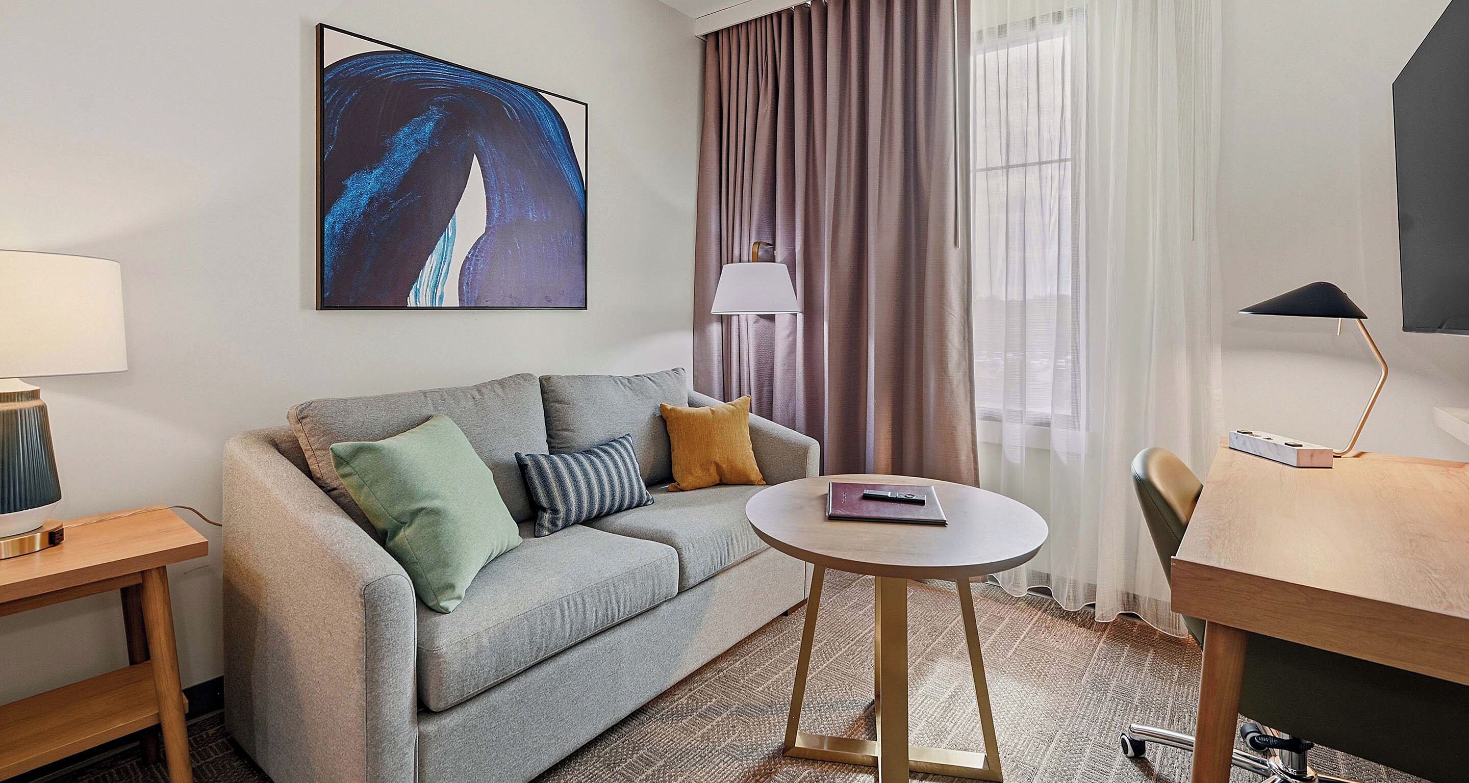 A hotel room with a sofa, a small round table, a lamp, a painting on the wall, and a view through the window.