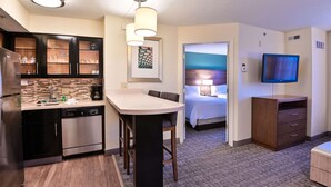 A modern hotel room with a kitchen area, a dining table, a TV, and a bedroom with a bed and nightstand.
