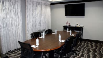 A conference room with a wooden table, black chairs, a flat-screen TV, a water dispenser, and a small bookshelf.