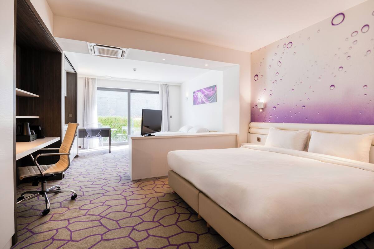 A modern hotel room with a large bed, a desk with a chair, a television, and a view of the outdoors.