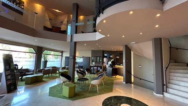 A modern hotel lobby with a high ceiling, multiple seating areas, a staircase, and a view of the street outside. - B&B HOTEL Mons Centre (Mons)