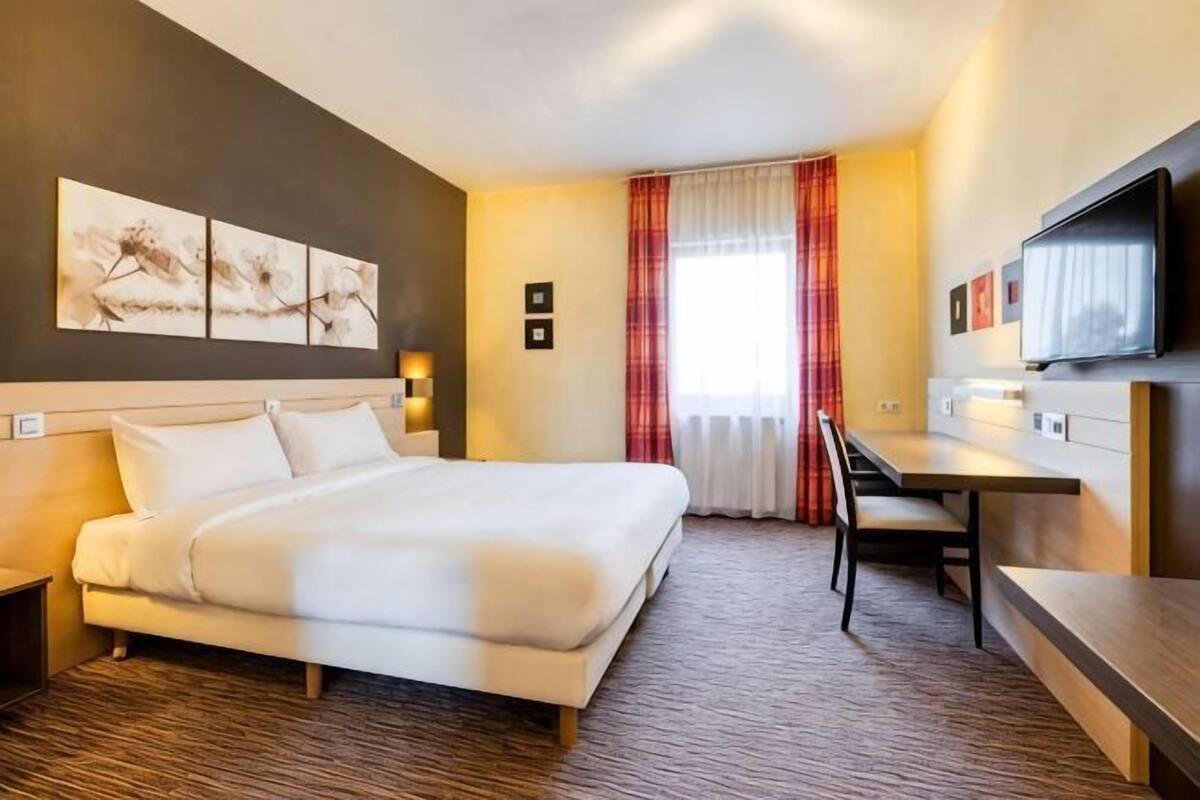 A hotel room with a bed, a desk, a chair, a TV, and a window with curtains.