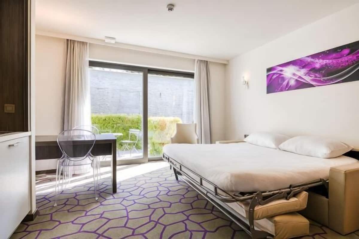A hotel room with a bed, a desk, a chair, a window with curtains, and a view of greenery.