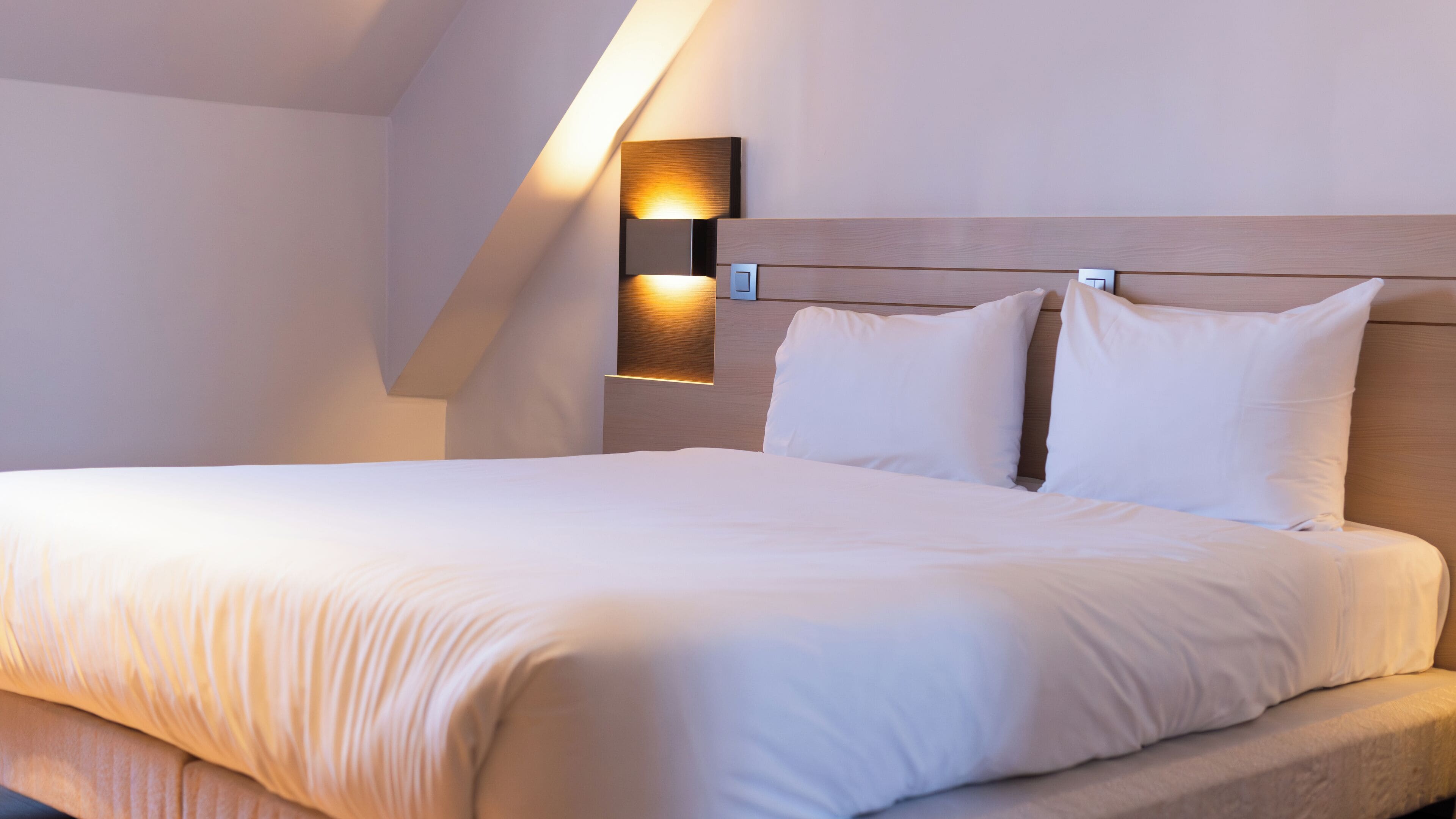 A neatly made bed with white linens in a modern hotel room with a sloped ceiling and a wall-mounted light fixture.