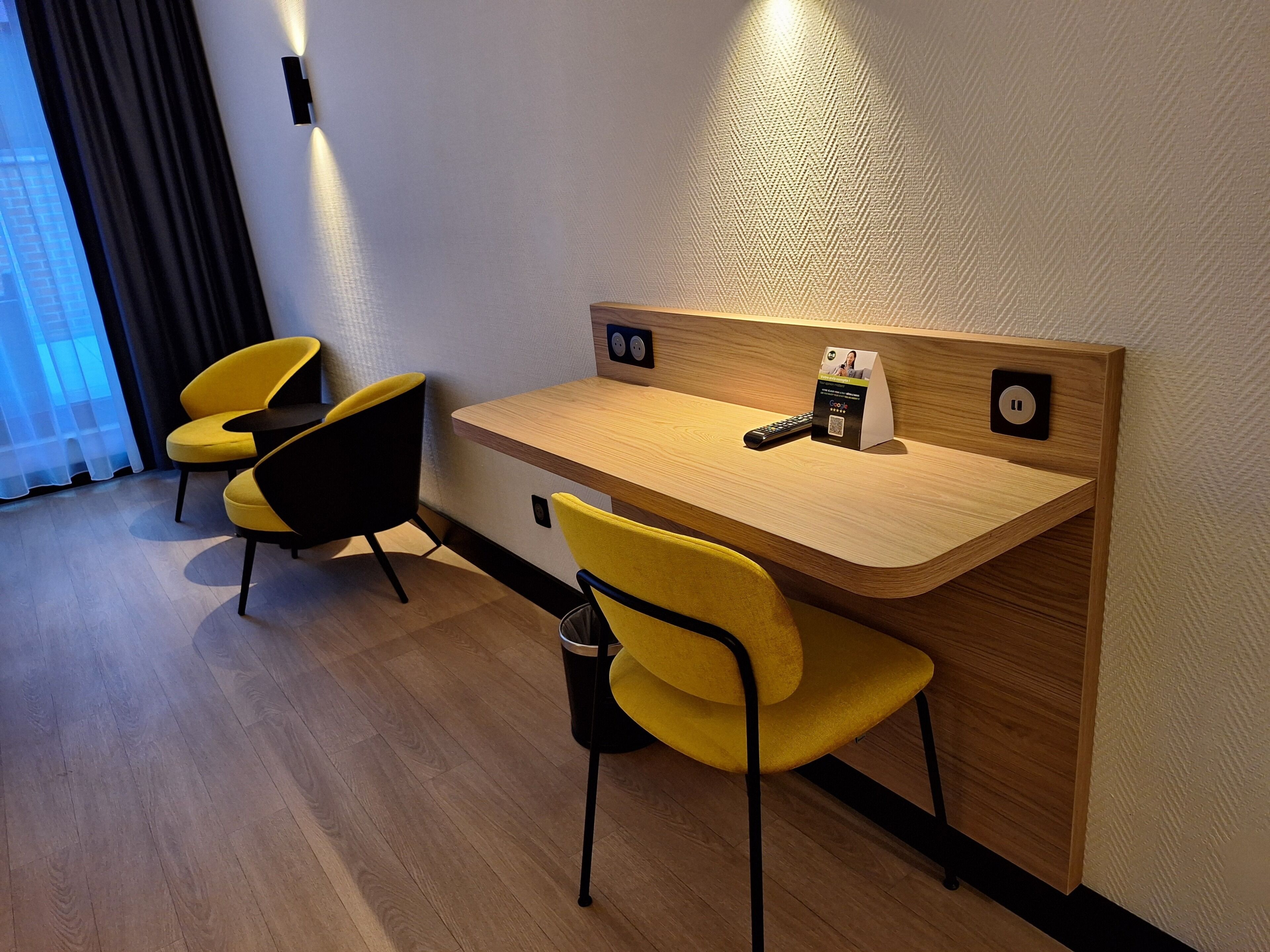 Habitación de hotel con un escritorio de madera, dos sillas amarillas, un control remoto y un cartel con un código QR.
