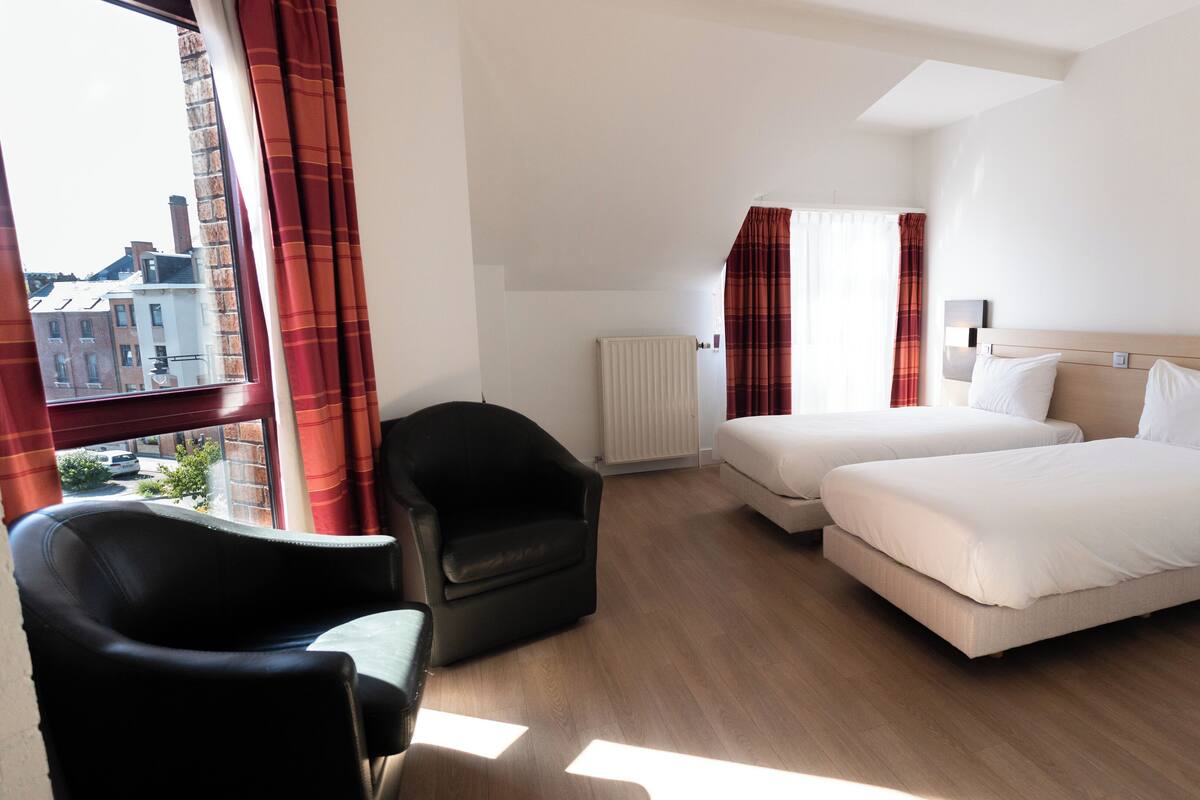 A hotel room with two beds, a chair, a radiator, and a window with curtains.