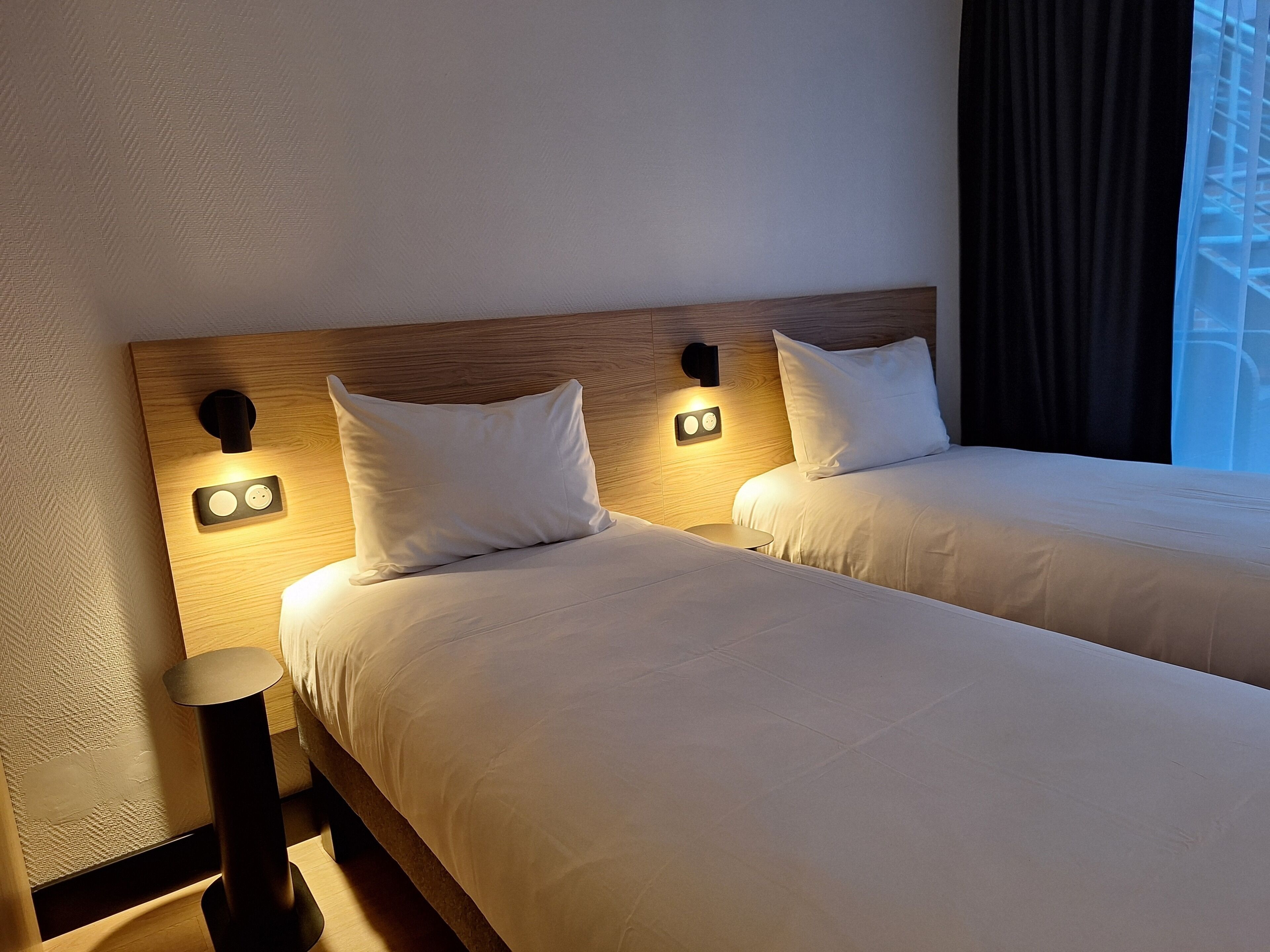 A hotel room with two beds, a wooden headboard, bedside lamps, and a window with a view.