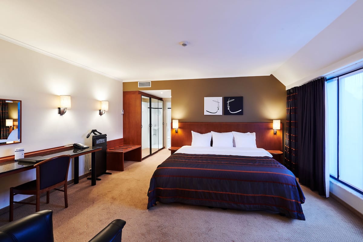 A hotel room with a large bed, a desk with a chair, a desk lamp, a telephone, and a mirror. There is a door leading to a bathroom and a window with curtains.