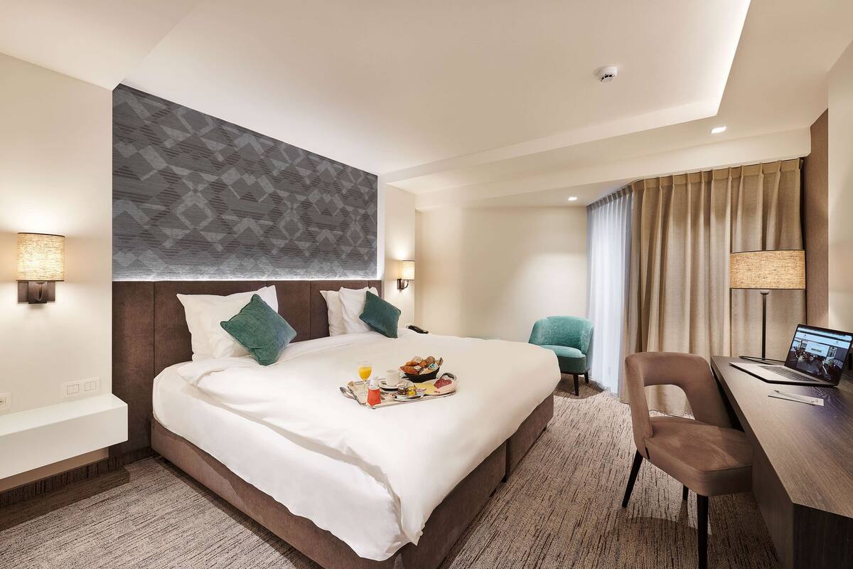 A modern hotel room with a large bed, a desk with a laptop, and a breakfast tray on the bed.