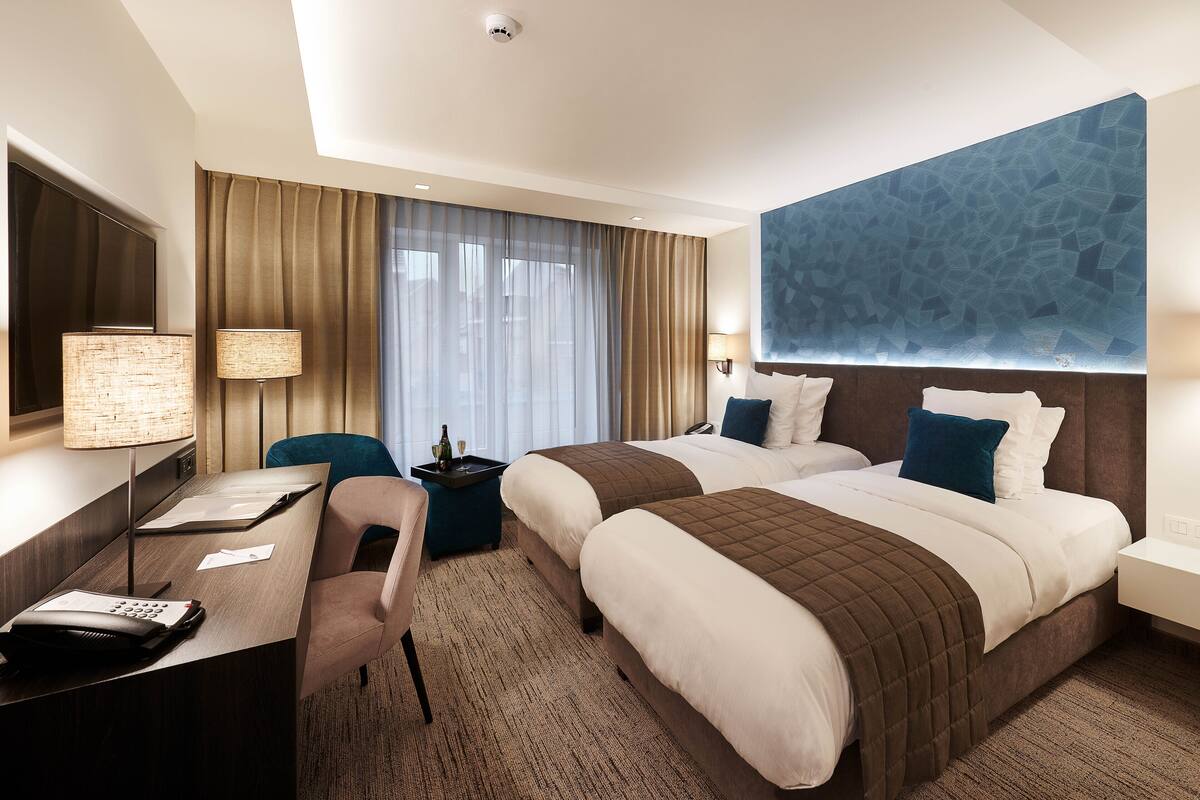 A hotel room with two beds, a desk with a telephone, a chair, a desk lamp, and a large abstract wall art piece.