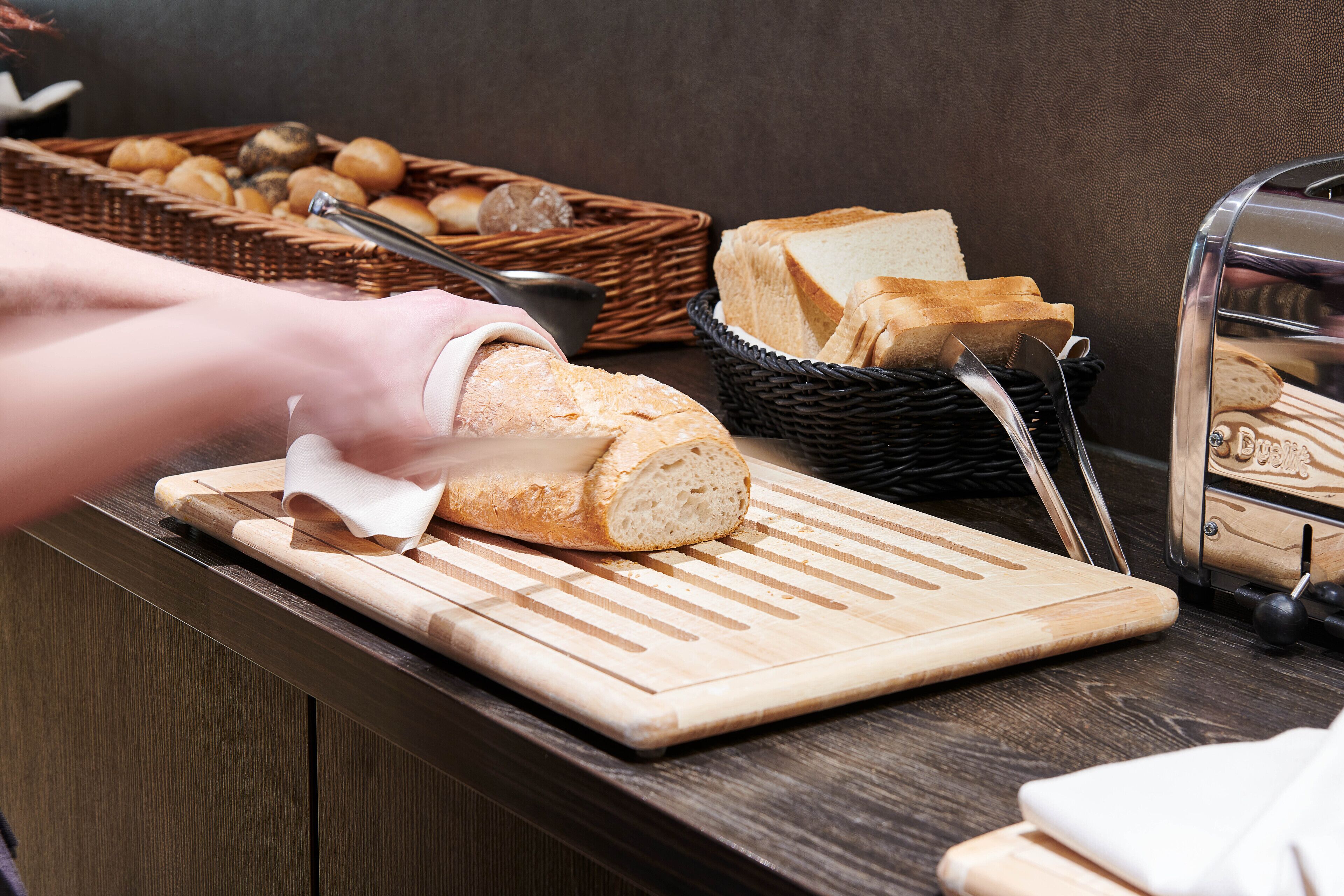 A kitchen counter with a loaf of bread being sliced, a basket of bread rolls, and a bread display case.