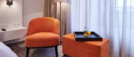 A hotel room with a single orange chair, a small ottoman with a tray of orange juice, a lamp, and a window with sheer curtains.