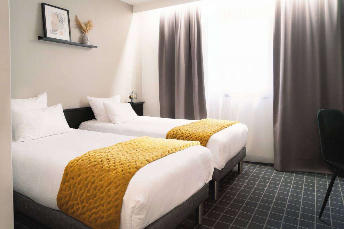 Ein Hotelzimmer mit zwei Betten, einem kleinen Tisch, einem Stuhl und einem Fenster mit Vorhängen.