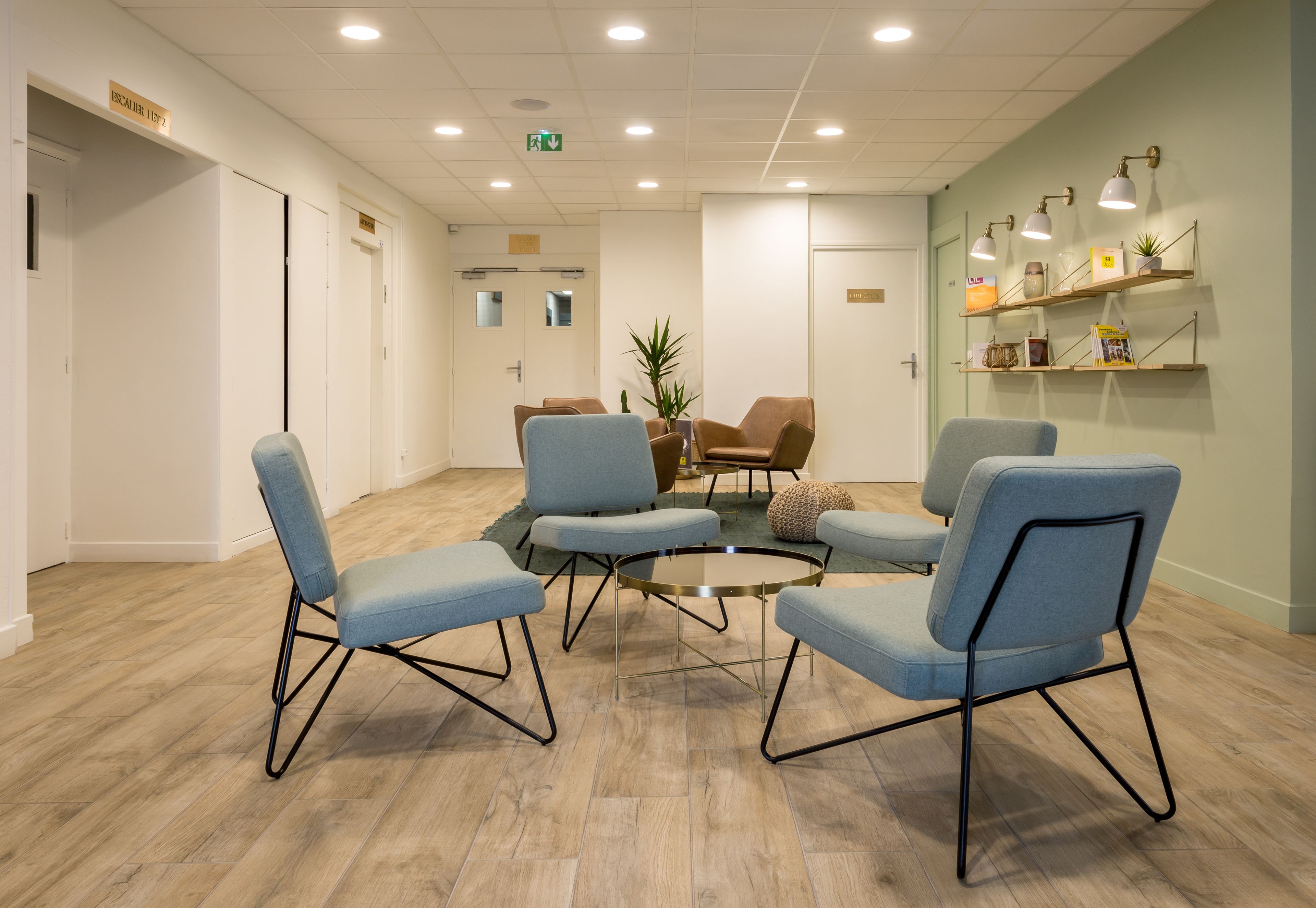 Un espace d’attente avec quatre chaises bleues disposées autour d’une table centrale, un plancher en bois et un mur vert agrémenté d’étagères.