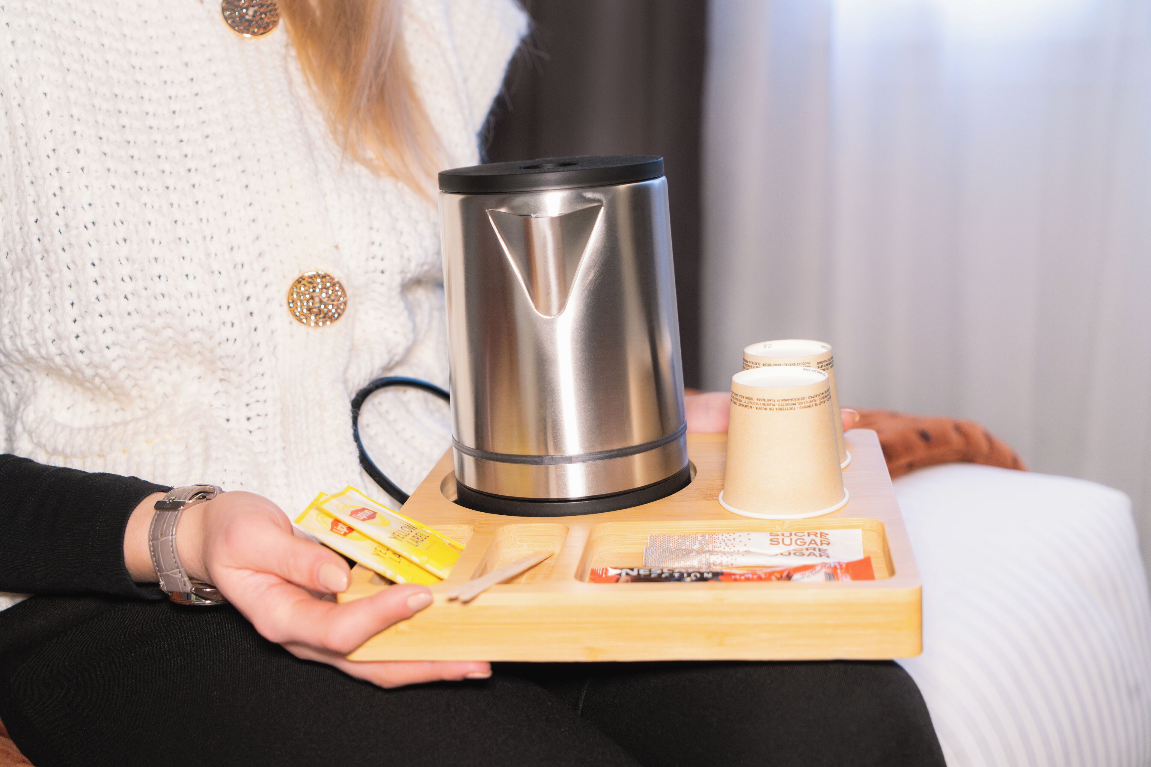 A person holding a tray with a stainless steel kettle, a candle, and some food items.