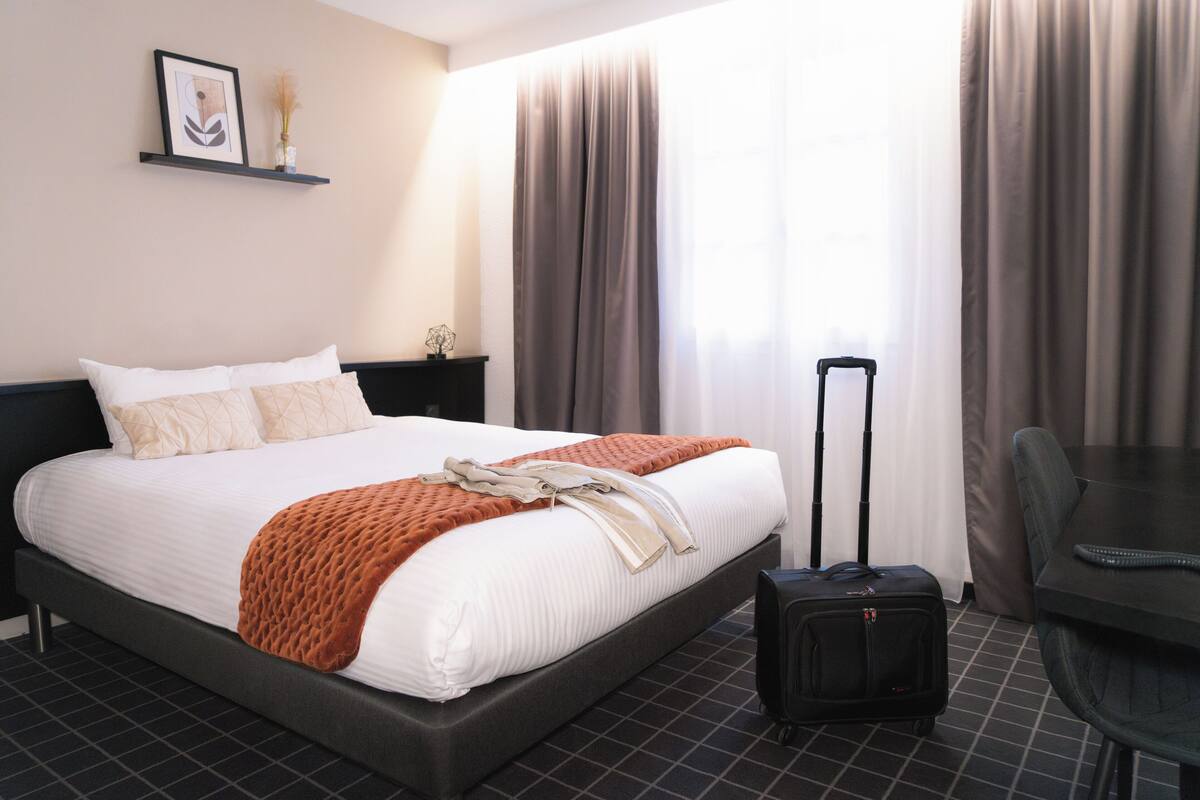 Ein Hotelzimmer mit Bett, Schreibtisch, Stuhl, Koffer und einem Fenster mit Vorhängen.
