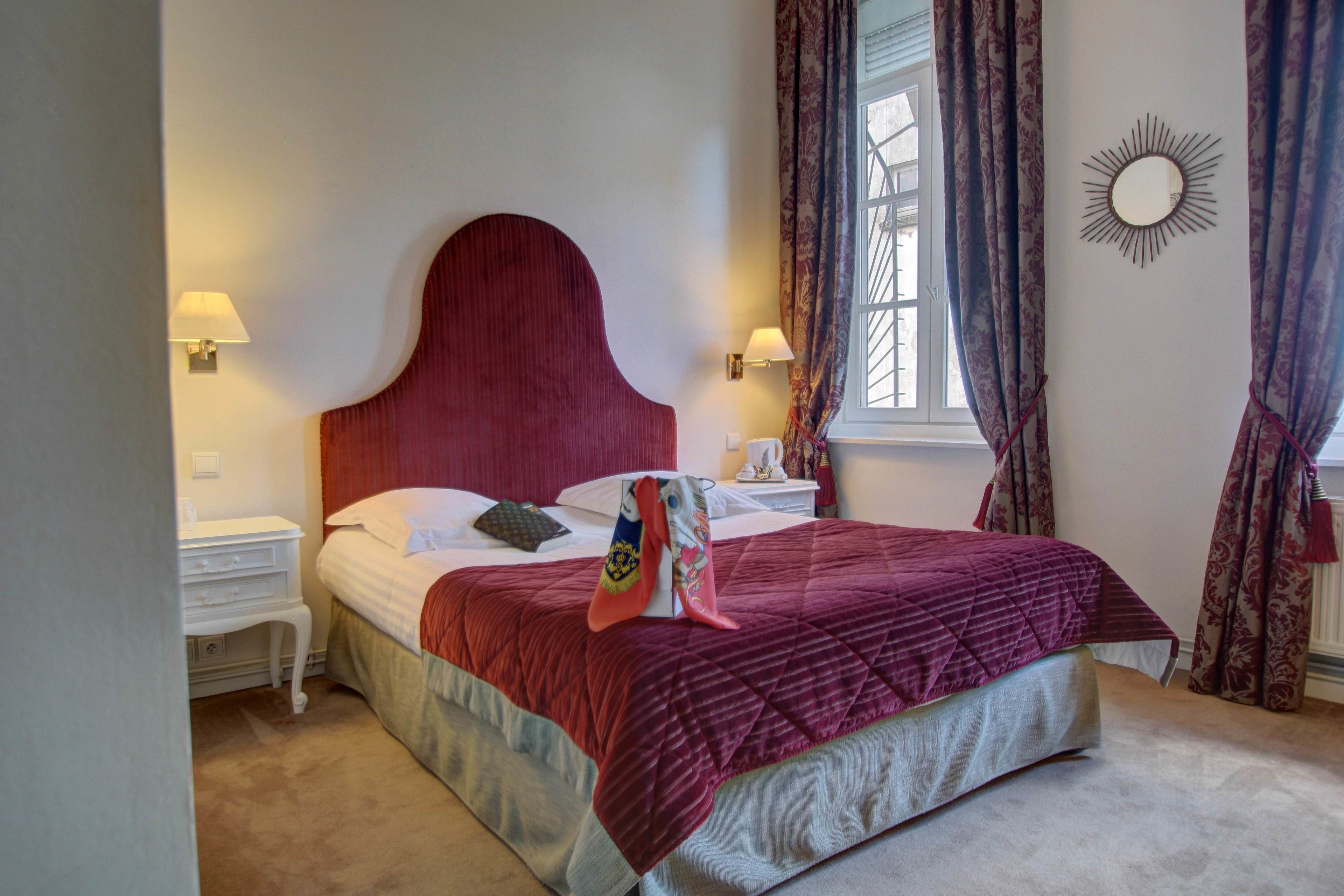Una cama bien hecha con cabecera roja, ropa de cama blanca y un cojín decorativo. Hay dos mesitas de noche con lámparas, una ventana con cortinas y un espejo montado en la pared.