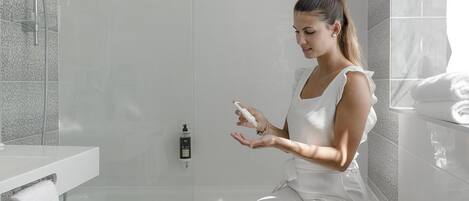 Une femme vêtue de blanc est assise sur une baignoire blanche dans une salle de bain moderne, équipée d’une douche, d’un porte-serviettes et d’un flacon de shampoing.