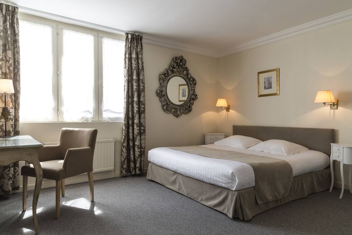 A hotel room with a bed, a chair, a desk, a window with curtains, a wall-mounted mirror, and two wall lamps.
