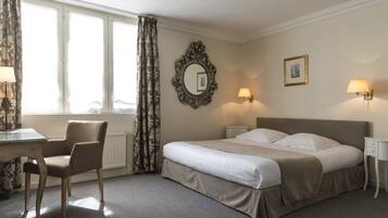 A hotel room with a bed, a chair, a desk, a window with curtains, a wall-mounted mirror, and two wall lamps.