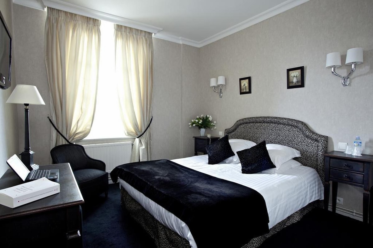 A hotel room with a large bed, a desk with a laptop, a chair, a nightstand with a lamp, and a window with curtains.